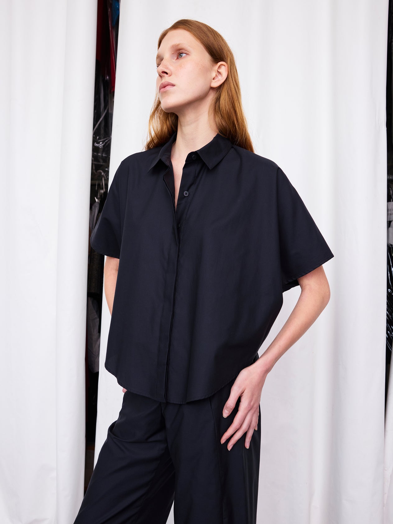 A person with long red hair, wearing the Zero + Maria Cornejo Circle Shirt and a white skirt, stands indoors with one arm raised behind their head. Potted plants and a black bench are visible in the background.