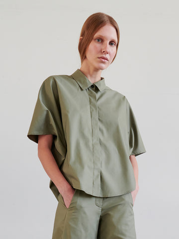 A person with light brown hair in a low ponytail wears the Circle Shirt by Zero + Maria Cornejo in olive green, paired with matching pants, against a plain light background.