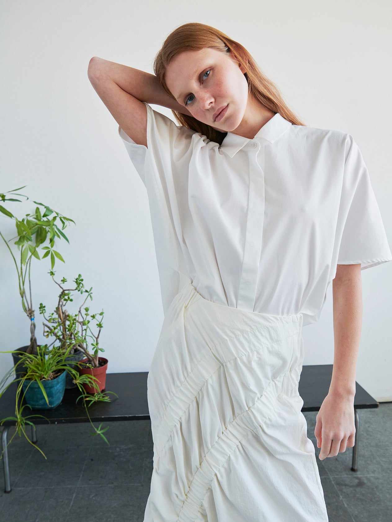A person with long red hair, wearing the Zero + Maria Cornejo Circle Shirt and a white skirt, stands indoors with one arm raised behind their head. Potted plants and a black bench are visible in the background.