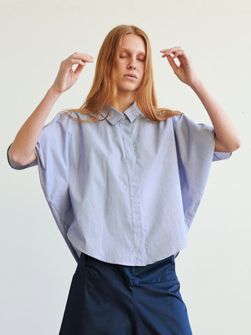 A person with long red hair wears the Zero + Maria Cornejo Circle Shirt, a loose striped cotton top with a curved hem, and dark pants, standing with eyes closed and hands raised against a plain light background.