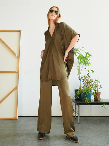 Confidently standing indoors near a bench with potted plants, a person wears sunglasses and the olive green Gigi Pant by Zero + Maria Cornejo, styled with a matching loose-fitting top featuring an elastic waist and wide-leg silhouette.