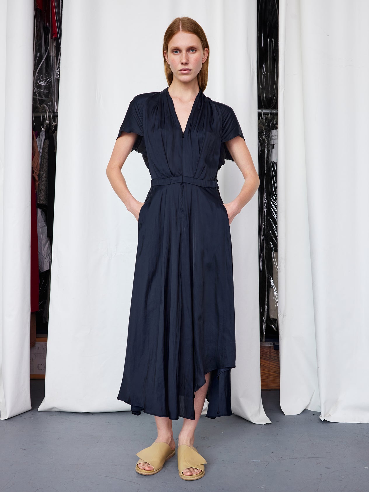 Indoors, a person wears the Zero + Maria Cornejo Maki Wave Dress—a dark blue, short-sleeve, wrap-style mid-length dress with pockets—paired with beige slip-on sandals, standing before white curtains.