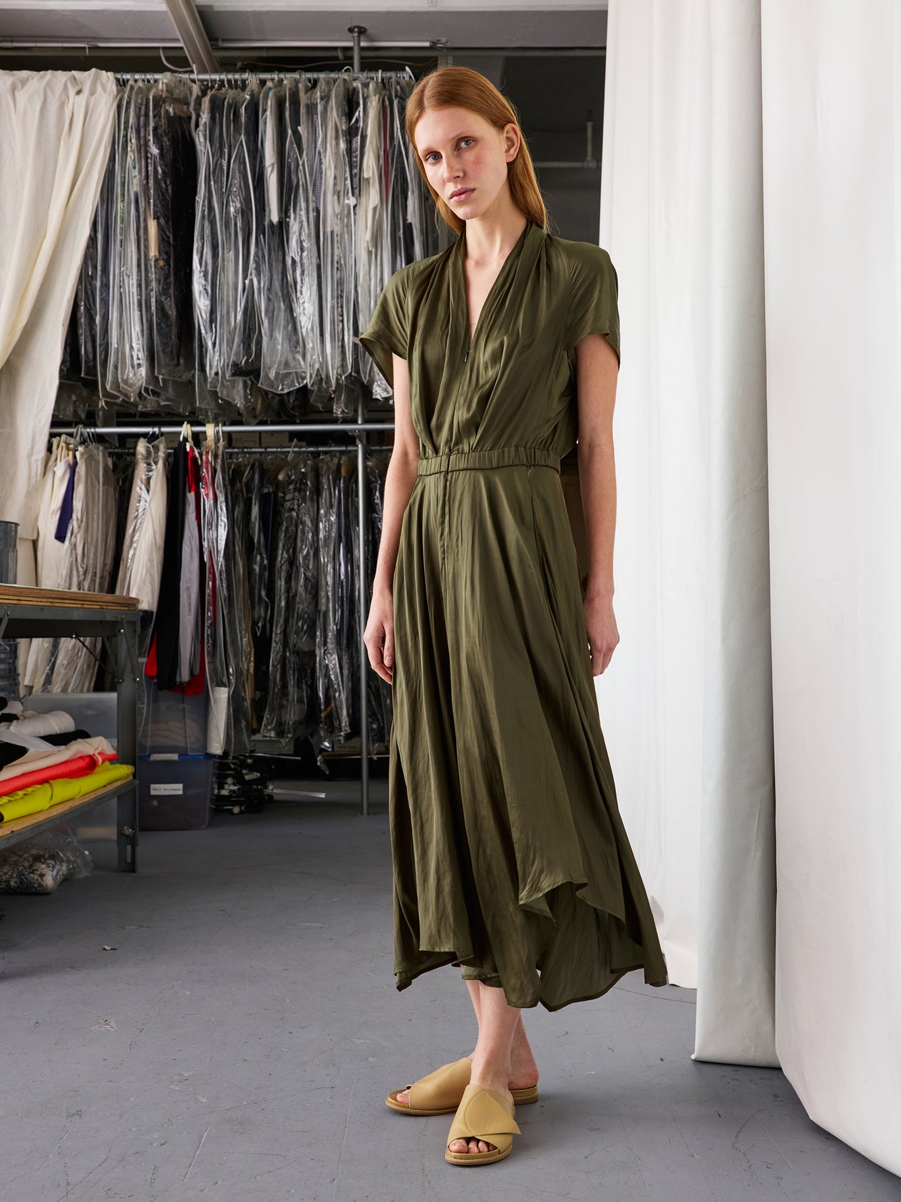 Indoors, a person wears the Zero + Maria Cornejo Maki Wave Dress—a dark blue, short-sleeve, wrap-style mid-length dress with pockets—paired with beige slip-on sandals, standing before white curtains.