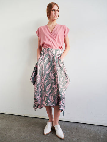 A woman with straight light brown hair stands against a plain wall, wearing Zero + Maria Cornejo’s Nio Skirt—an abstract pink and gray jacquard leaf-patterned high-waisted sustainable skirt—with a pink wrap top and white pointed-toe shoes.