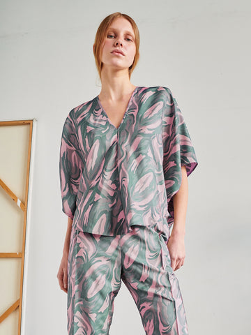 A person stands indoors in the Circle Top by Zero + Maria Cornejo, a loose recycled polyester set with pink and green abstract swirls. The minimal background includes a partial wooden frame on the left.