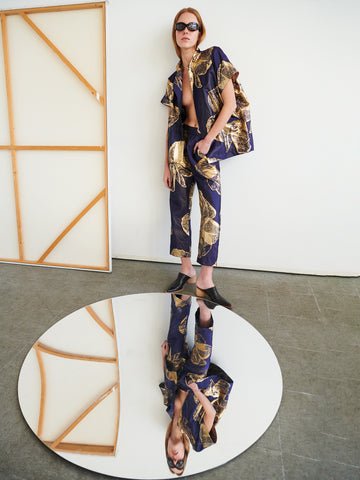 Wearing Zero + Maria Cornejo's Eko Pant and sunglasses, a person stands on concrete beside a large round mirror, which reflects their full look along with part of the plain wall and framed canvas behind them.