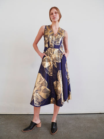 A woman stands against a white wall, wearing the Zero + Maria Cornejo Sleeveless Wave Dress, a navy blue midi dress with a gold floral jacquard pattern. Her hands are in her pockets and she pairs it with black low-heeled slip-on shoes.