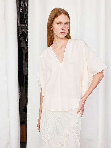 A woman with long red hair stands before white curtains, wearing the lightweight Circle Top by Zero + Maria Cornejo and a ruffled white skirt, looking directly at the camera with a neutral expression.