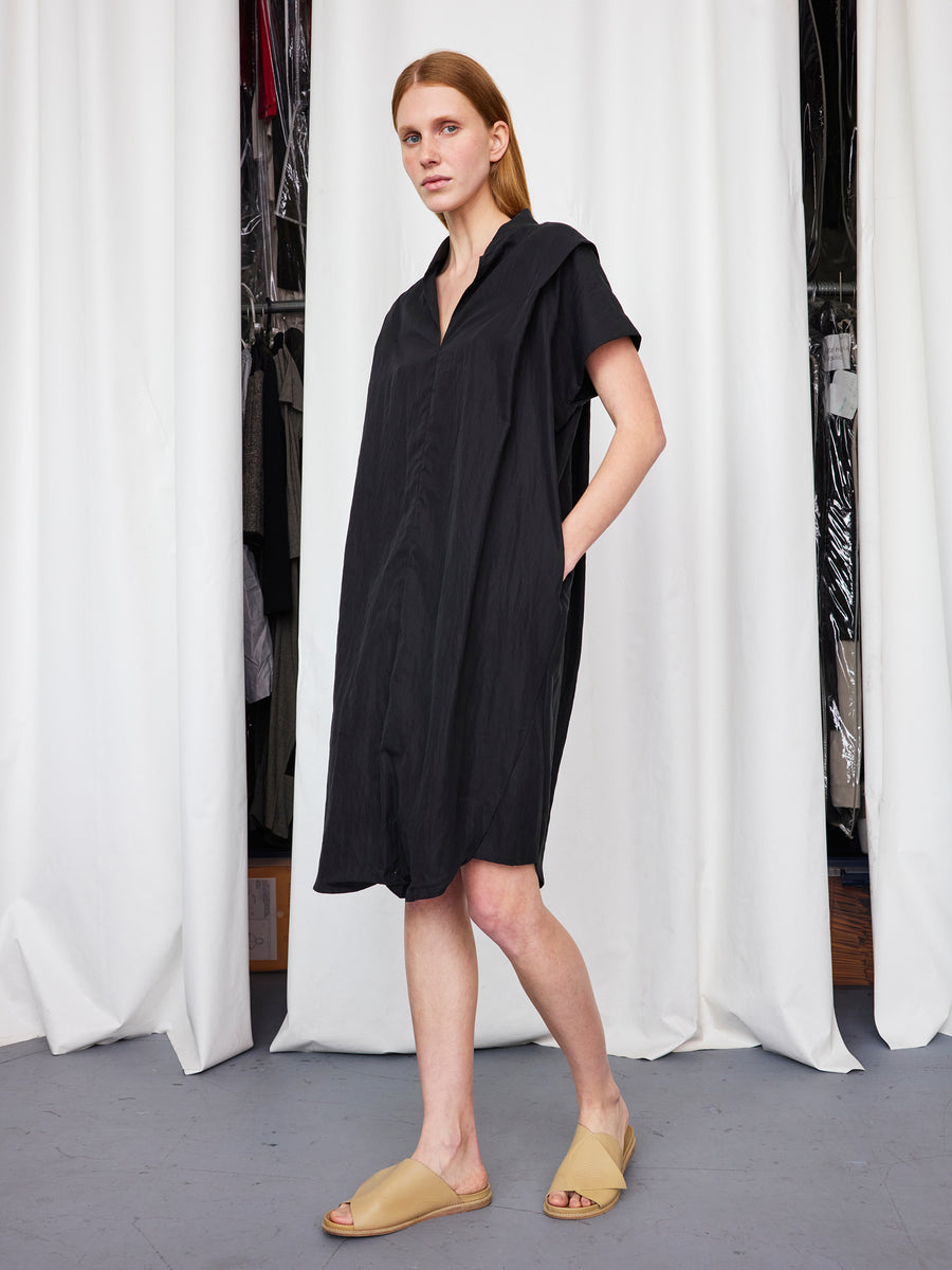A woman with straight, light brown hair wears the Zero + Maria Cornejo Pleat Circe Dress, a loose-fitting, knee-length TENCEL Lyocell style, and beige slide sandals while standing before white curtains with clothes in the background.