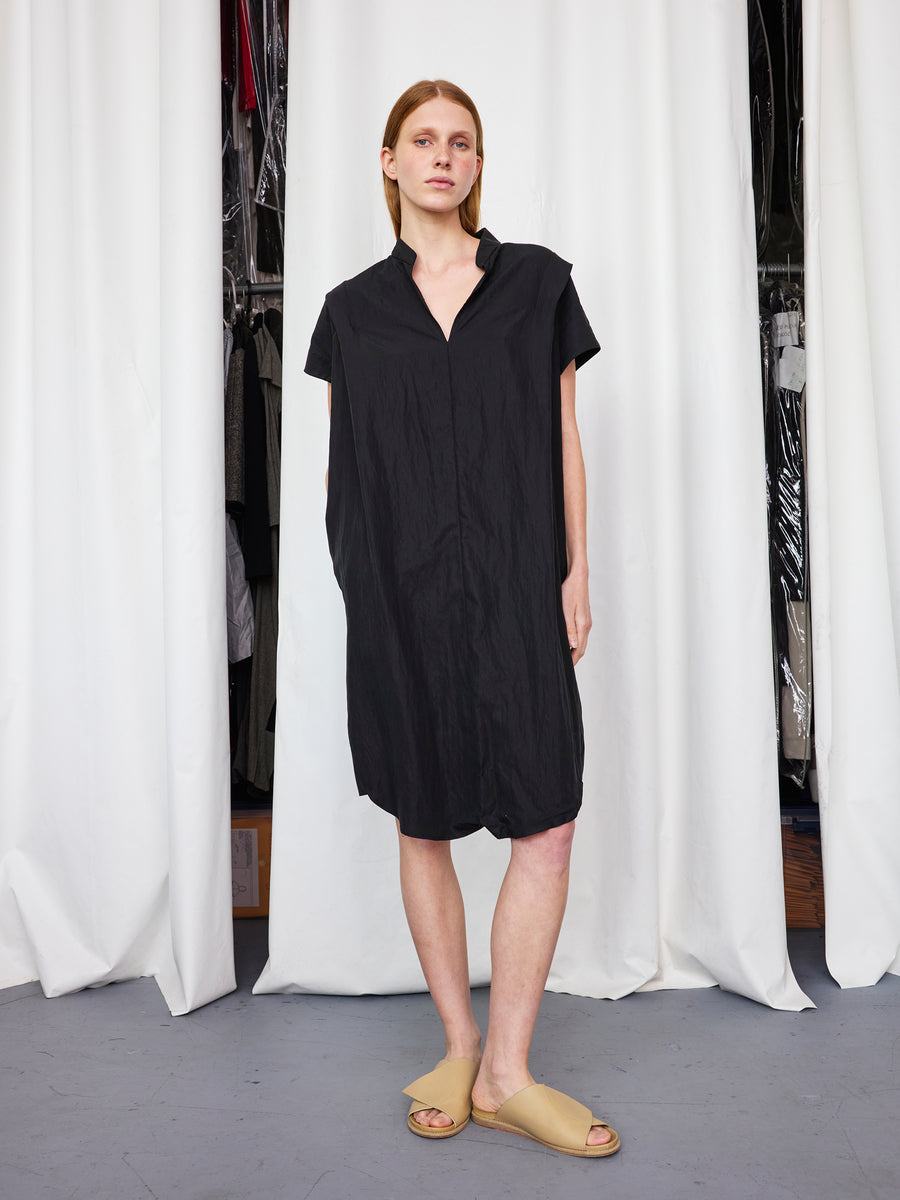 A person with light skin and straight, light brown hair stands by white curtains, wearing the Zero + Maria Cornejo Pleat Circe Dress and beige slide sandals. Hanging clothes are partially visible in the background.