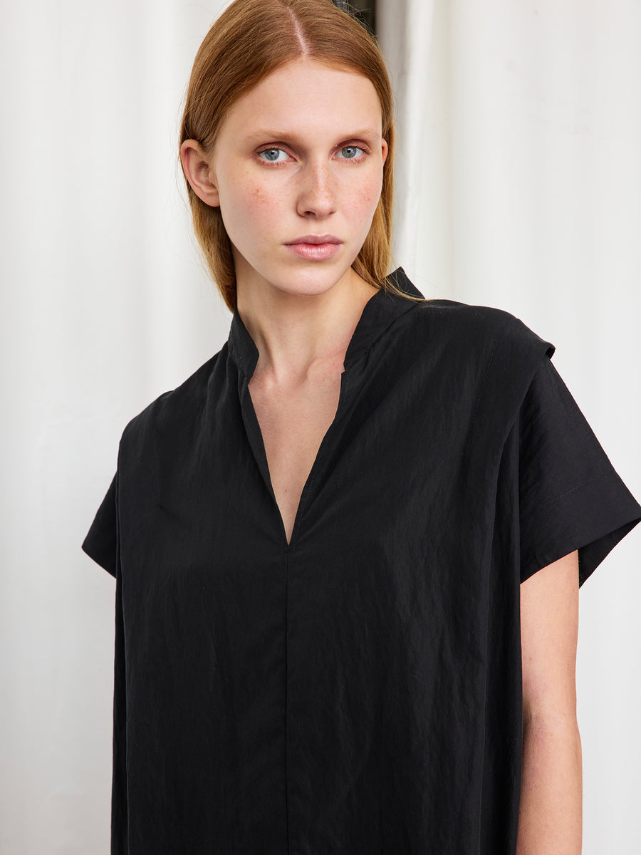 A woman with straight, light brown hair and blue eyes stands before a white curtain, wearing Zero + Maria Cornejo's Pleat Circe Dress—a loose-fitting, knee-length style with v-neck and mandarin collar—looking calmly at the camera.