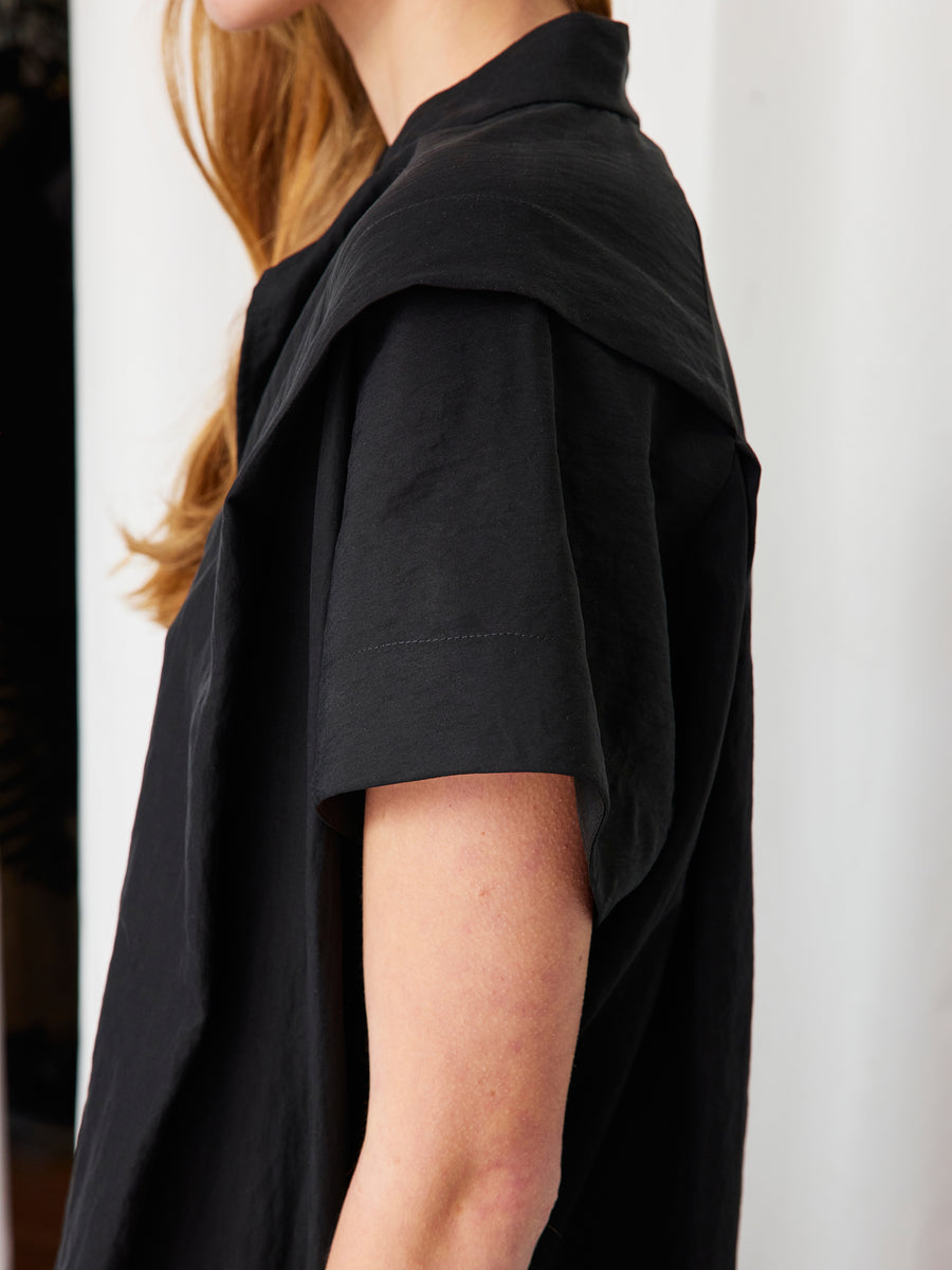 A woman with long, wavy light brown hair models the Zero + Maria Cornejo Pleat Circe Dress in black TENCEL Lyocell. She stands sideways against a white background, showing only the upper half of her body.
