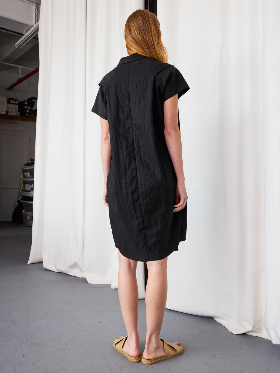 A person with long hair stands facing away, wearing the Zero + Maria Cornejo Pleat Circe Dress and tan slip-on sandals, in a room with white curtains and a gray floor.