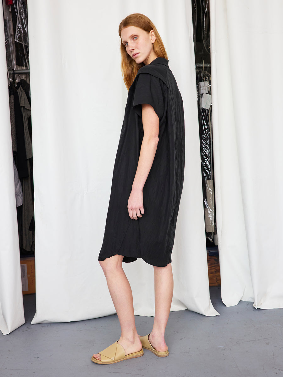 A person with long light brown hair models the Zero + Maria Cornejo Pleat Circe Dress—loose-fitting, knee-length—paired with beige sandals, standing sideways before white curtains and a clothing rack.