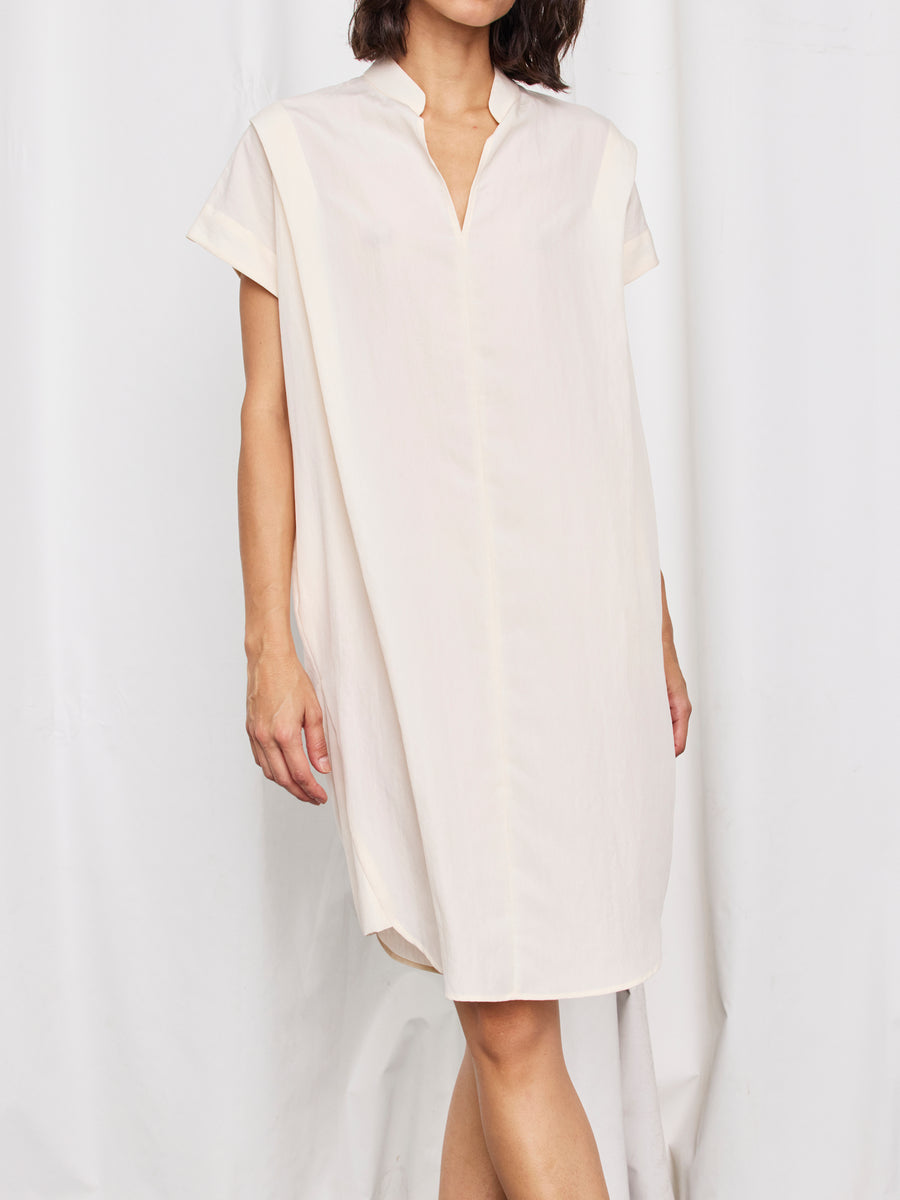 A woman in the Zero + Maria Cornejo Pleat Circe Dress—a loose, lightweight, knee-length piece with short sleeves, collar, and V-neck made from sustainable fabric—stands against a white curtain; her face is partially hidden.