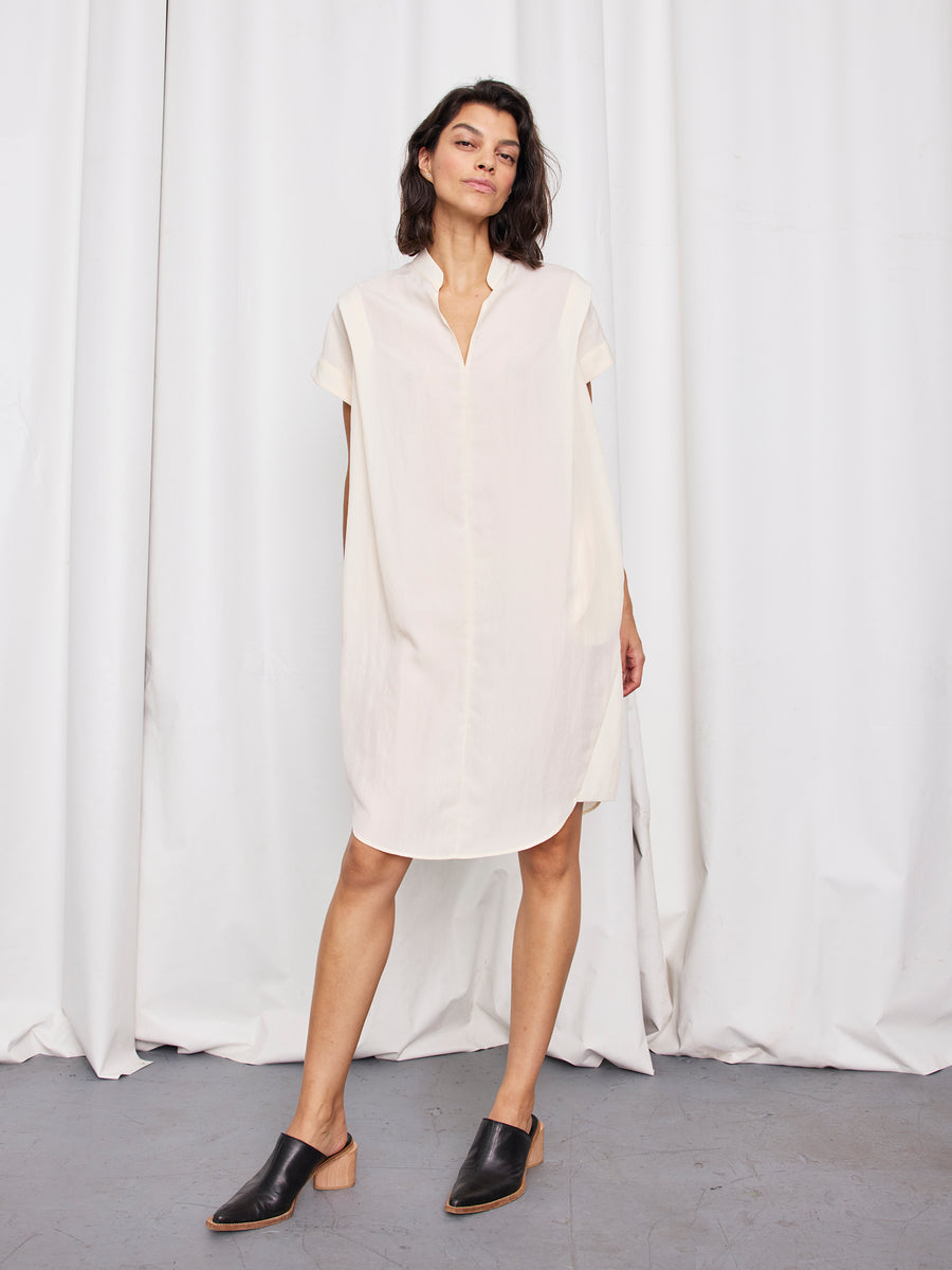 A woman stands confidently in front of white curtains, wearing the Zero + Maria Cornejo Pleat Circe Dress—a lightweight, knee-length TENCEL Lyocell design—paired with black low-heel slip-on mules made in New York.