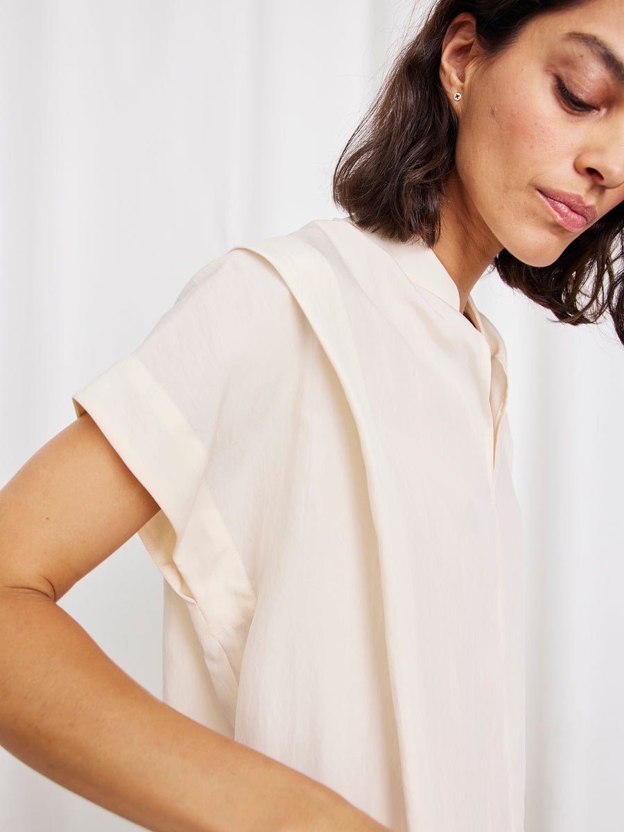 A woman with short dark hair wears the Pleat Circe Dress by Zero + Maria Cornejo, a loose, cream TENCEL Lyocell piece, standing against a plain light background and turned slightly to the side with her face partly visible.