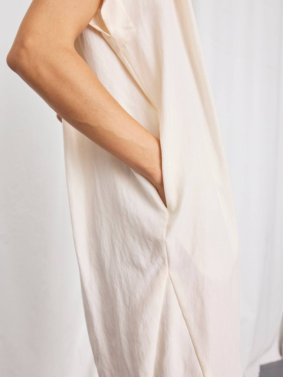 Someone stands against a light background, one hand in their pocket, wearing the Pleat Circe Dress by Zero + Maria Cornejo—a loose, sleeveless, cream-colored dress made from TENCEL Lyocell.