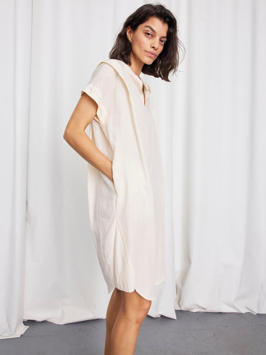 A woman with shoulder-length dark hair stands sideways, modeling the Zero + Maria Cornejo Pleat Circe Dress—a short-sleeve, light cream, knee-length TENCEL Lyocell dress with pockets—posing before white curtains.