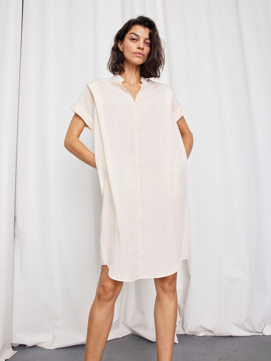 A woman stands facing forward with hands in pockets, wearing the Pleat Circe Dress by Zero + Maria Cornejo—a loose, short-sleeved, lightweight knee-length style in light beige—against a plain white fabric background.