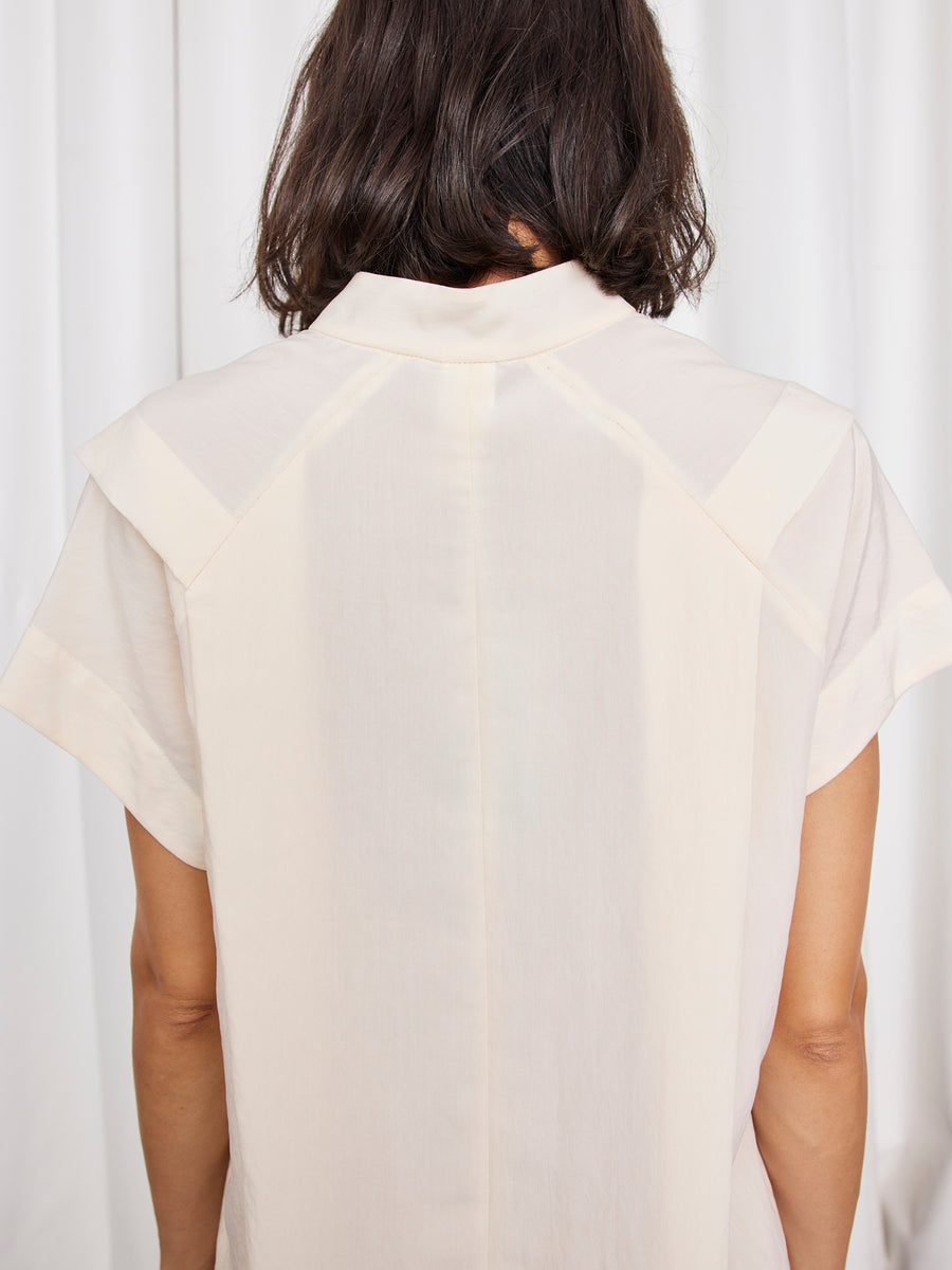 A person with dark, shoulder-length hair is seen from behind wearing the Pleat Circe Dress by Zero + Maria Cornejo—a light TENCEL Lyocell dress with short sleeves and a collar—set against a backdrop of white curtains.