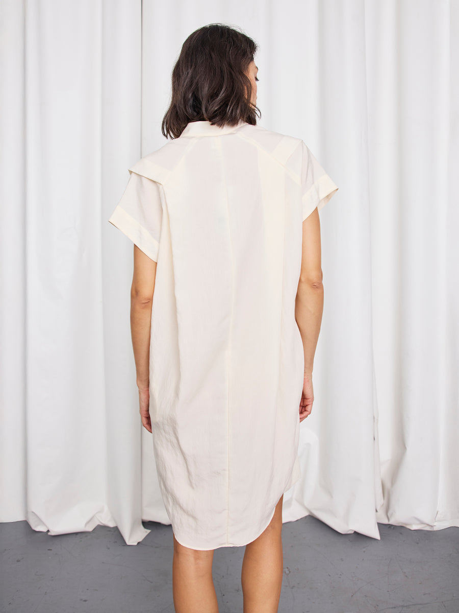 A person with shoulder-length dark hair, seen from behind facing white curtains, wears the knee-length Pleat Circe Dress by Zero + Maria Cornejo, crafted from sustainable fabric.
