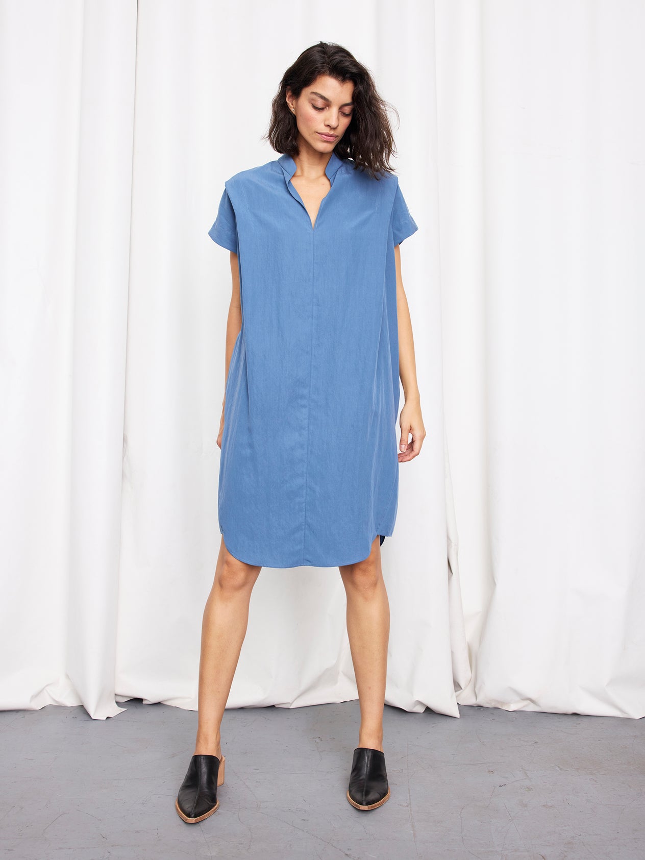 A woman with straight, light brown hair stands before white curtains, wearing the Pleat Circe Dress by Zero + Maria Cornejo—a loose, knee-length TENCEL Lyocell dress with a V-neck— and looks at the camera with a neutral expression.
