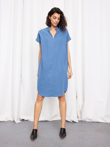A woman stands before white curtains wearing the Pleat Circe Dress by Zero + Maria Cornejo, a lightweight, knee-length style with short sleeves, paired with black mule shoes; her hands rest at her sides and feet are slightly apart.