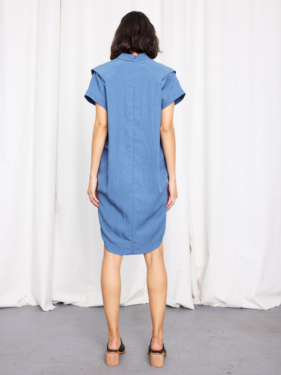 A woman faces away from the camera, wearing the Zero + Maria Cornejo Pleat Circe Dress in blue with black sandals, standing on a gray floor before white curtains.