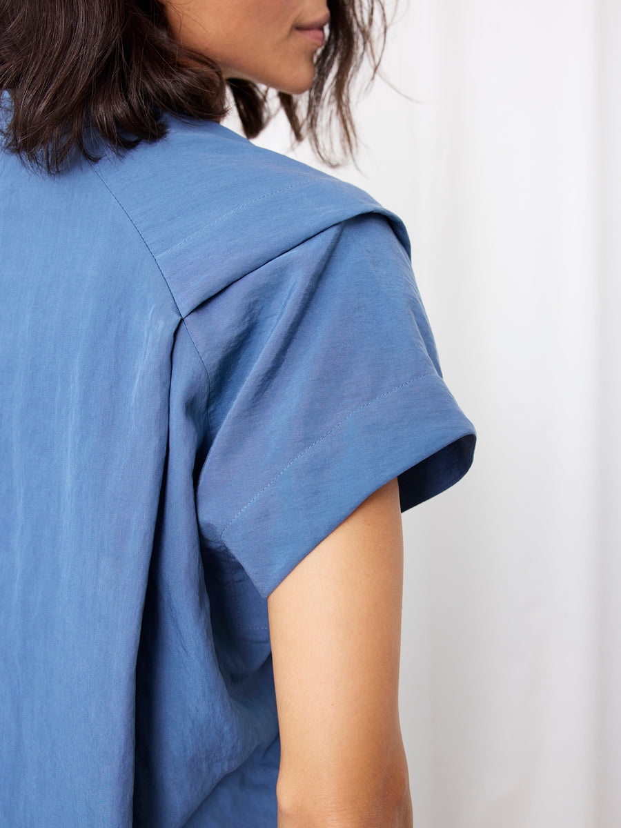 A person with shoulder-length dark hair models the short-sleeved, blue Pleat Circe Dress by Zero + Maria Cornejo. The image captures their upper body from the side, highlighting the dress’s sleeve against a plain, light background.