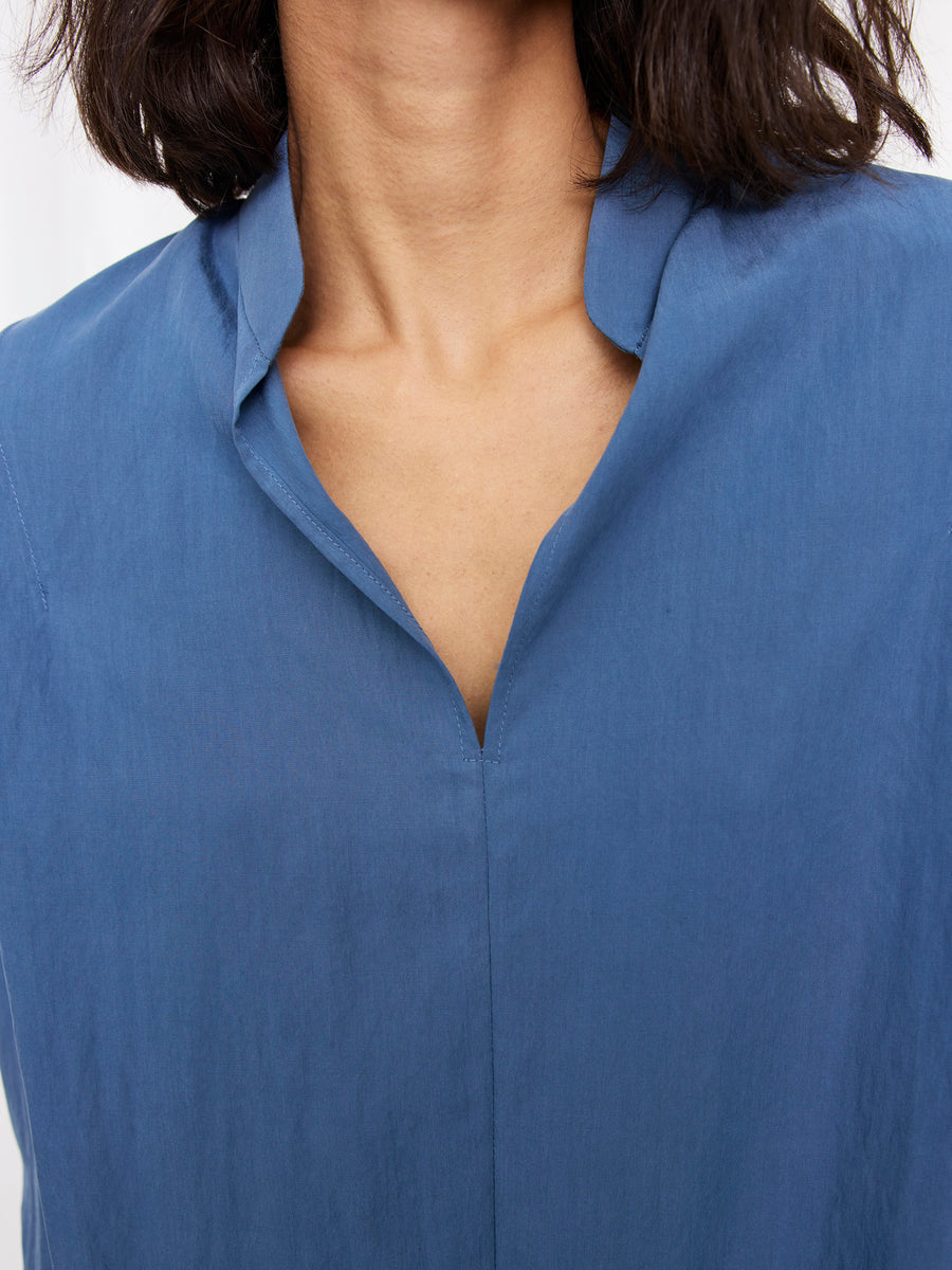 A person wearing the Pleat Circe Dress by Zero + Maria Cornejo, featuring a blue TENCEL Lyocell fabric and V-shaped neckline; only the lower face, neck, and upper chest are visible.