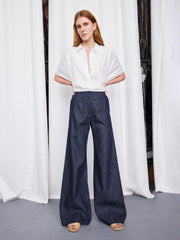 A woman with straight red hair stands before white curtains, wearing a short-sleeve white button-up shirt, beige open-toe sandals, and the Zero + Maria Cornejo Wide Eda Pant.