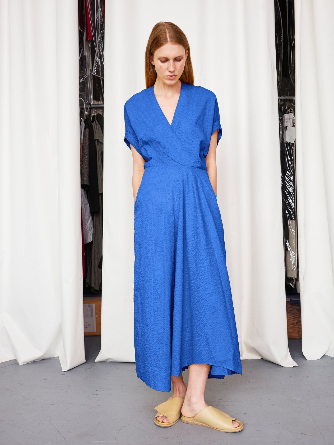 A woman stands before white curtains in the Long Aki Wave Dress by Zero + Maria Cornejo—a bright blue, lightweight textured cotton dress with a wrap-effect v-neck, asymmetrical skirt, and pockets. She looks down, hands in her pockets.