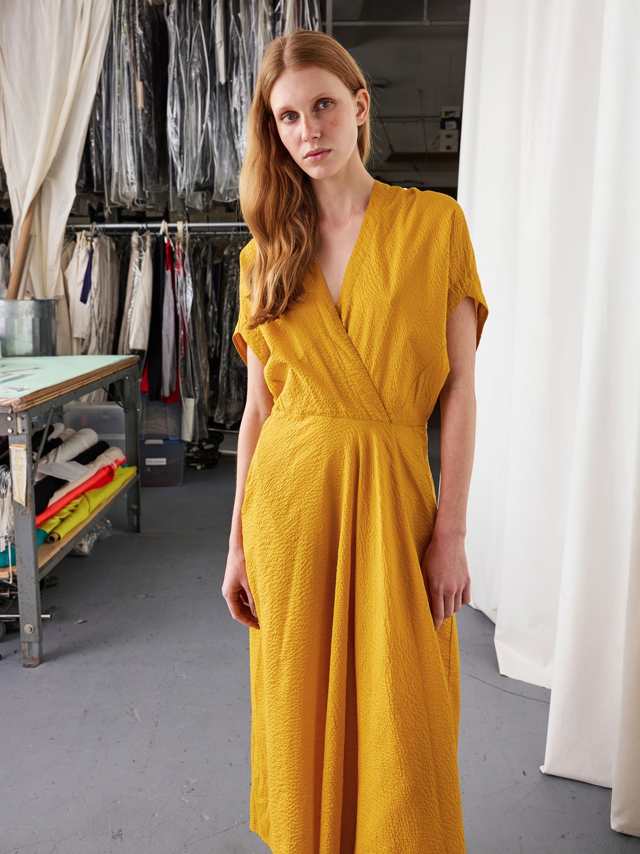 A woman stands before white curtains in the Long Aki Wave Dress by Zero + Maria Cornejo—a bright blue, lightweight textured cotton dress with a wrap-effect v-neck, asymmetrical skirt, and pockets. She looks down, hands in her pockets.
