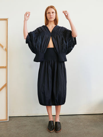 A person with straight auburn hair stands indoors against a white wall, wearing the Evia Shrug in dark navy woven voile by Zero + Maria Cornejo with a matching smocked skirt and black slip-on shoes. Their arms are raised slightly.
