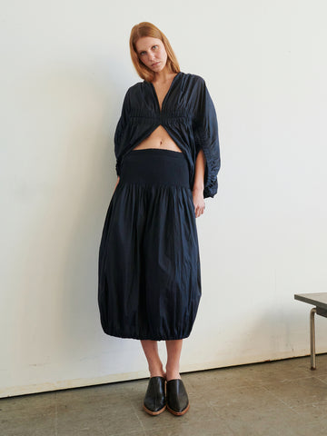 A person stands indoors against a white wall, wearing a dark blue long-sleeve top with a front cutout and the Eva Bubble Skirt by Zero + Maria Cornejo, featuring subtle smocked gathering. They have straight light brown hair and black slip-on shoes.