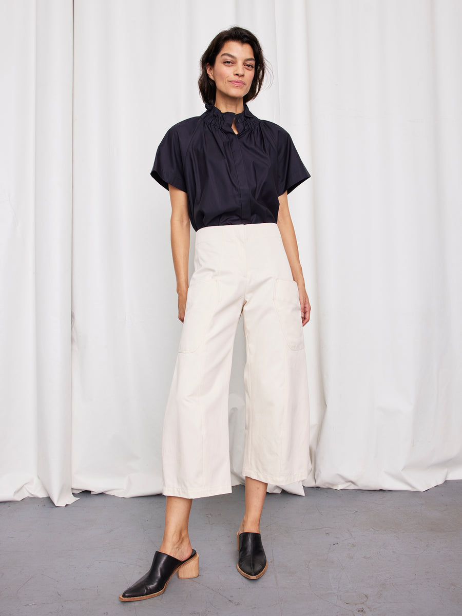 A woman with short dark hair and a relaxed expression stands by white curtains, wearing a short-sleeve navy blouse and Zero + Maria Cornejo Curved Eda Culotte pants, styled with black mule heels.