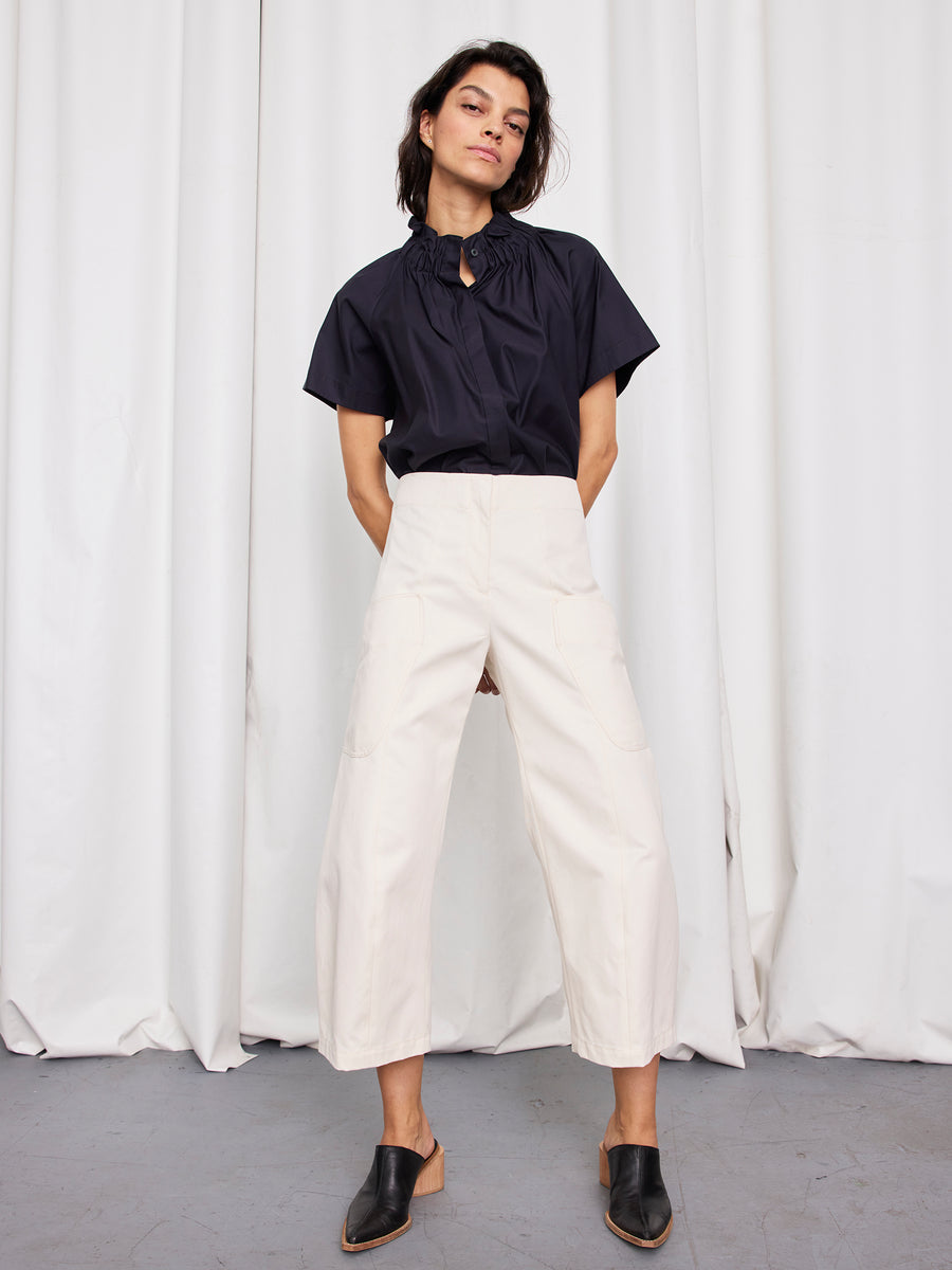 A person stands before white curtains wearing a short-sleeve black blouse, the Zero + Maria Cornejo Curved Eda Culotte with a wide leg, and black slip-on shoes. Their hands rest in their pockets as they confidently face the camera.