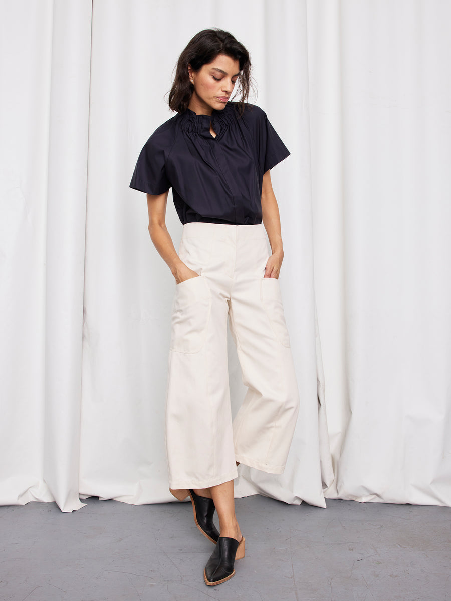 A woman stands before white curtains wearing a short-sleeved black blouse, Zero + Maria Cornejo Curved Eda Culotte, and black mule shoes. With her hands in her pockets, she gazes slightly downward.