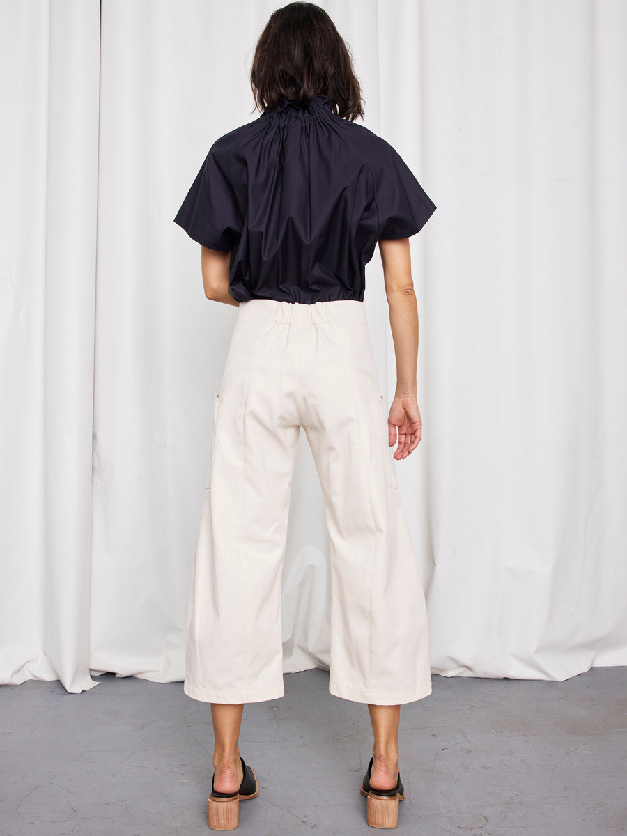 A person stands facing away from the camera in front of white curtains, wearing a short-sleeve black top, black sandals with block heels, and Zero + Maria Cornejo’s Curved Eda Culotte.