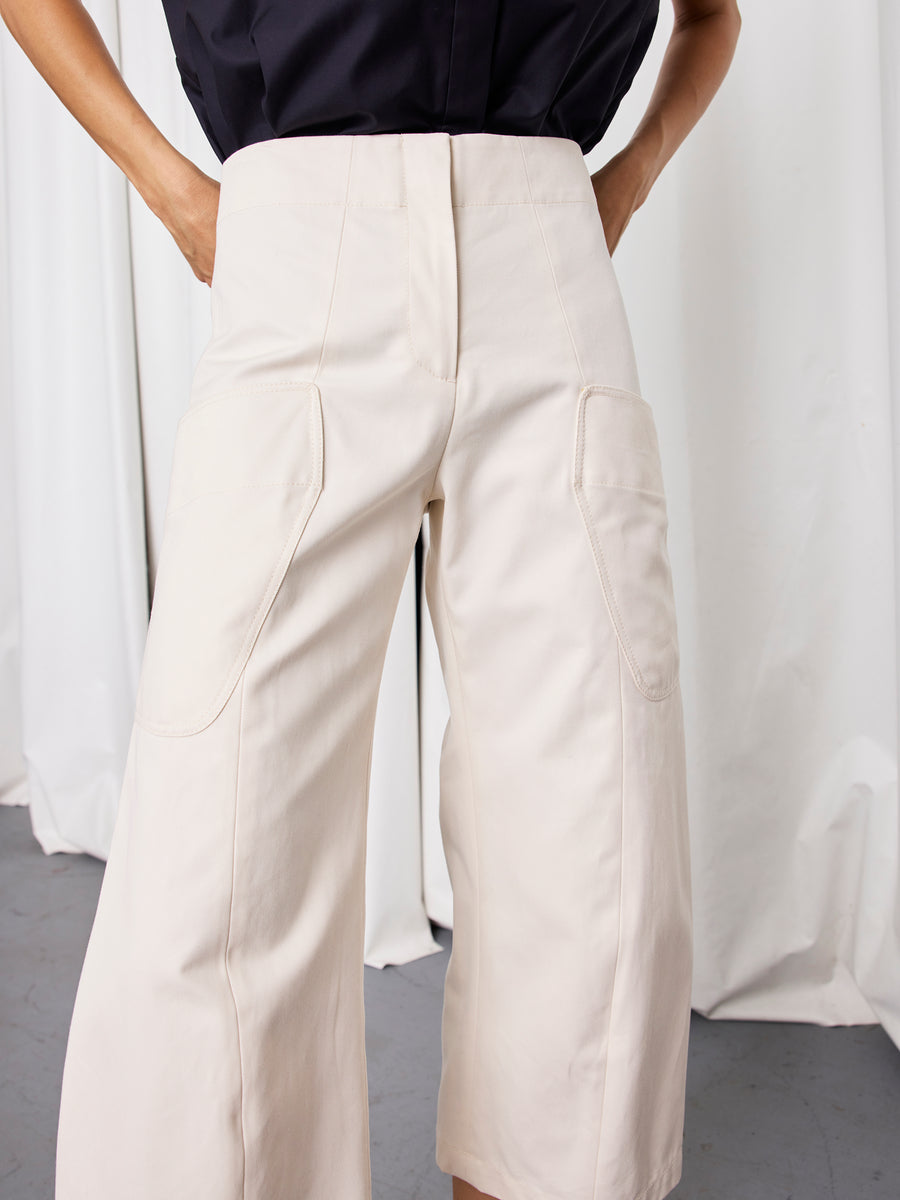 A person stands with hands on hips in front of white curtains, wearing the Zero + Maria Cornejo Curved Eda Culotte—high-waisted, wide leg cotton twill pants with large front pockets—and a tucked-in black top.
