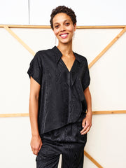 A person with short curly hair, smiling in front of a light background with wooden cross beams, wears the black Fin Gaban Shirt by Zero + Maria Cornejo and matching pants.