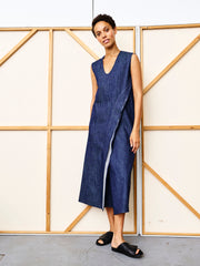 A person with short curly hair stands before a beige and wood-paneled backdrop, wearing the Zero + Maria Cornejo Long Twisted Tank Dress in indigo organic denim and black slide sandals.