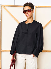 A woman in dark sunglasses and the black Long-Sleeved Akeo Shirt by Zero + Maria Cornejo stands before a beige and wood panel background, wearing light beige pants and holding a burgundy handbag.