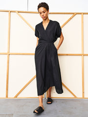 A person stands indoors before a wooden and white panel, wearing the black Long Aki Wave Dress by Zero + Maria Cornejo with black slide sandals, one hand in their pocket and looking down.