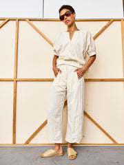 Wearing sunglasses, a loose cream top, Zero + Maria Cornejo’s Aissa Cargo Pant, and beige slide sandals, a person stands with hands in pockets before a wooden-framed off-white backdrop.