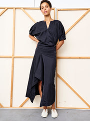 A woman stands before a wooden frame wall, wearing a loose black V-neck top with puffed sleeves, the black Ero Skirt by Zero + Maria Cornejo, and white slip-on shoes. She looks at the camera with her hands in her pockets.