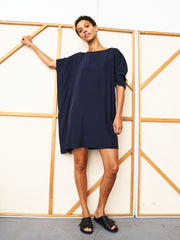 A person with short hair poses against a wall in the Zero + Maria Cornejo Long-Sleeved Contra Dress, a navy above-knee shift style with loose sleeves, styled with black slide sandals and minimal jewelry.