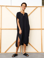 A person stands smiling in front of a beige and wooden panel wall, wearing the Rae Caftan in black by Zero + Maria Cornejo, styled with black slide sandals and minimal jewelry.