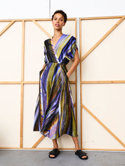 A person poses confidently before a wooden panel wall, wearing the Long Aki Wave Dress by Zero + Maria Cornejo—a vibrant silk charmeuse design with pockets and black slide sandals. This standout piece is made in New York.